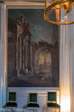 <center>Grande antichambre.</center>Paire de tableaux : architecture et ruines. Paltronieri Pietro. 1720-1740. Paire de toiles peintes représentant des caprices architecturaux en ruine envahis par la végétation où sont disposés ça et là quelques groupes de figures. La première toile représente un groupe de trois personnages (deux femmes et un enfant) dans un paysage d'architecture grandiose. Une colonnade en marbre veiné bleu encadrant un escalier côtoie d'immenses colonnes cannelées à chapiteaux corinthiens. En arrière-plan, de hauts murs en pierre de taille aux coloris roses sont ornés de médaillons et de frises à denticules et percés d'arcades brisées à panneaux moulurés.Sur la droite, trois vaches paissent près d'un escalier. Sous l'une des arcades se trouve une fontaine à laquelle s'accoudent un homme et un enfant. Près d'eux, un enfant joue avec une canne et tient son chapeau devant un immense socle portant un vase, sur un fond de paysage rocheux.