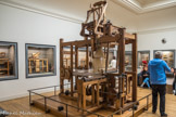 <center>Musée des Arts et Métiers.</center> Métier à tisser les façonnés de Vaucanson. Jacques Vaucanson, célèbre dans toutes les cours d’Europe pour ses automates, est nommé en juin 1741 inspecteur des manufactures de soie. il commence alors une tournée d’inspection des établissements et imagine des machines qui améliorent grâce à des dispositifs mécaniques le moulinage de la soie ou le tissage des tissus façonnés.
Cames, manivelle et vis sans fin.
Le métier combine plusieurs recherches de Vaucanson : la programmation, la reproduction du mouvement humain et un système ingénieux Bile navette. Cette dernière, entièrement métallique, est saisie alternativement par des bras munis de pinces, fixés chacun sur un chariot de bois. La mécanique, le chasse- navette et le battant sont entraînés par des cames. Le tissu s’enroule régulièrement grâce à une vis sans fin.
Un automate à tisser.
Véritable automate à tisser, le métier est entièrement mis en mouvement par une simple manivelle et transforme ainsi radicalement les gestes du tisseur. En 1747, Le Mercure de France rapporte : « On voit sur le métier l’étoffe se fabriquer sans aucun secours humain, c’est-à-dire la chaîne s’ouvrir, la navette jeter la trame, le battant frapper l’