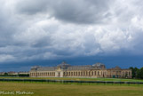 <center>Chantilly. Les grandes écuries. </center> Au premier plan, l'Hippodrome. Construit à dessein le long de la Pelouse, face à la forêt et donnant aujourd'hui sur le champ de course, le monument actuel s'étend en longueur sur 186 mètres de façade, divisée en deux ailes encadrées, réunies par un avant-corps octogonal, et cantonnées de deux pavillons aux extrémités. Le dôme s'élève jusqu'à 28 mètres de hauteur, couronné par la réplique de la statue équestre, 