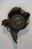 Anita Molinero. Sans titre, 2000.
Plastique (polypropylène)
Musée d’Art Moderne, Paris, France.
En 2000, Anita Molinero habite à Marseille, dans le quartier de la Belle de Mai. Une manifestation éclate, et un groupe de jeunes met le feu aux poubelles du quartier. Pour elle, ces containers fondus sont de véritables sculptures. C’est un « choc esthétique ».
Il s’agit de sa première poubelle, un bas-relief façonné « par les humeurs et les colères de la rue », désormais considérée comme un objet « signature » de sa pratique. Depuis cet événement, le mobilier urbain a pris une place à part entière dans sa sculpture. « Tout ce style ornemental que l’on voit a été fait par des racailles qui y ont foutu le feu. J’ai trouvé ça magnifique. Je récupérerai le terme de « style » comme il est employé par les jeunes. Elle a encore du style. C’est aussi un style ornemental presque des années 1930. Je l’ai déboulonnée, je l’ai récupérée, et j’y ai ajouté ce trou central, qui est un trou extrêmement expressionniste et qui est presque la représentation de la colère, du cri. »