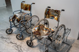 Sans titre (El cochecito), 2009-2014. Fauteuils roulants, parking à vélo, inox miroir
Courtesy de l’artiste et de la Galerie Christophe Gaillard, Paris, France
Anita Molinero a réalisé la série des chaises roulantes en pensant à un personnage du film La petite voiture (El Cochecito, 1960) de Marco Ferreri. Dans l’Espagne soumise à la dictature franquiste, une personne âgée rêve de pouvoir conduire une voiture. Son fauteuil roulant apparaît comme un outil paradoxal de liberté et d’émancipation. L’artiste le transforme en un bel objet rutilant. Elle remplace le cuir des assises et des dos par de l’aluminium très épais chauffé au chalumeau pour obtenir ces reflets moirés. Puis, elle a tordu le métal pour donner une impression de mouvement. Cependant, elle suspend leur mobilité en les fixant dans un parking à vélo. Ces objets d’un quotidien hors norme nous arrêtent par leur charge émotionnelle et politique. « Ils nous sidèrent et nous obligent à nous fixer. Ils s’opposent à la fluidité, à la rapidité. Par rapport à l’unité de la sculpture, leurs mesures ergonomiques sont justes.
Et puis la vieillesse, c’est probablement le prochain âge créatif et obscène. »