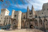 <center>Palais des Papes d'Avignon </center>Le Palais des papes vu de la Place du Palais. La construction du palais des Papes est l'œuvre principalement de deux papes bâtisseurs : Benoît XII, qui fait construire le premier palais pontifical (dit Palais vieux), et Clément VI, qui fait édifier de nouvelles extensions (dit Palais neuf). Benoît XII, confie la direction du chantier à son compatriote du comté de Foix, l’architecte Pierre Poisson. Dès le début de son pontificat, Clément VI achève la tour de Trouillas, ajoute une nouvelle tour des cuisines et fait construire la Tour de la Garde-Robe, accolée à la Tour du Pape. Par la suite, l’architecte Jean de Louvres, originaire de l’Ile-de-France, acquiert le statut de maître des œuvres et supervise toute l’organisation du chantier des nouvelles extensions, qui mobilisera en moyenne 600 hommes dans un contexte pourtant troublé par la guerre de Cent Ans et la peste noire.